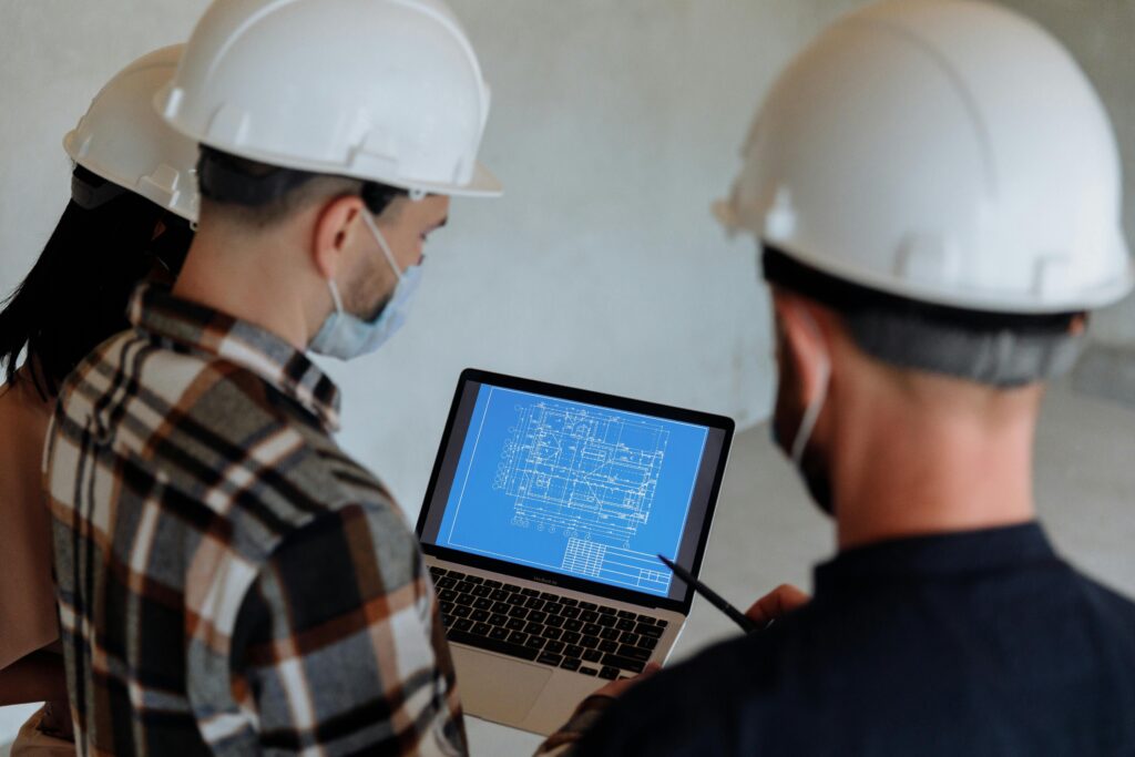 pexels-photo-8482865-8482865 Engineers wearing hard hats and masks review a construction blueprint on a laptop indoors.